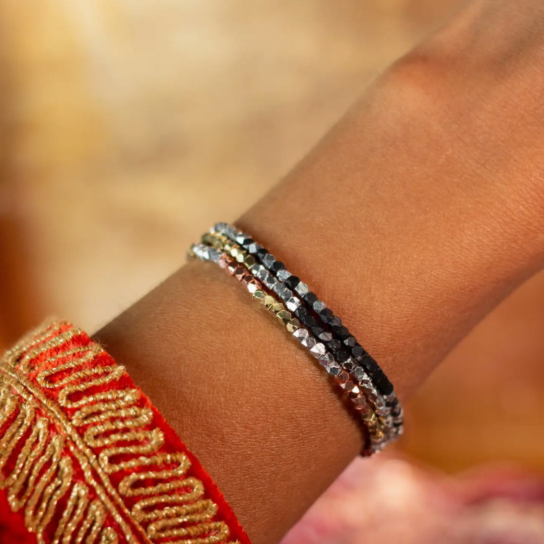 Photograph of a dark-skinned wrist adorned with three thin beaded bracelets featuring black, rose gold, and silver faceted beads, next to a red garment with intricate gold embroidery