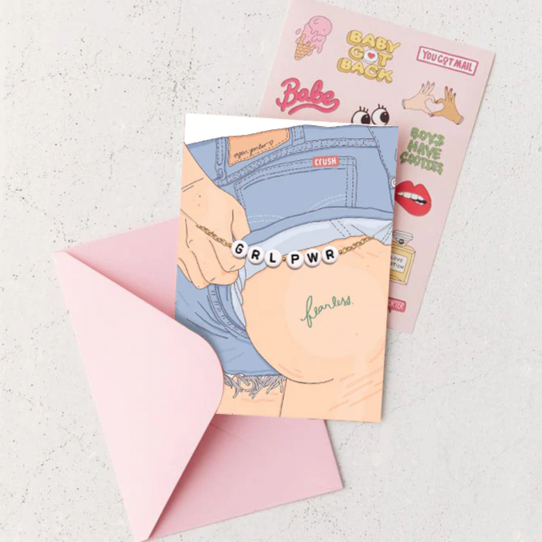 A cartoon greeting card depicting a person in denim shorts with a 'fearless' tattoo and a 'GRL PWR' bead chain, resting near a partially open pink envelope and a sticker sheet featuring colorful designs like 'BABY GOT BACK' and a melting ice cream cone, all on a light grey background