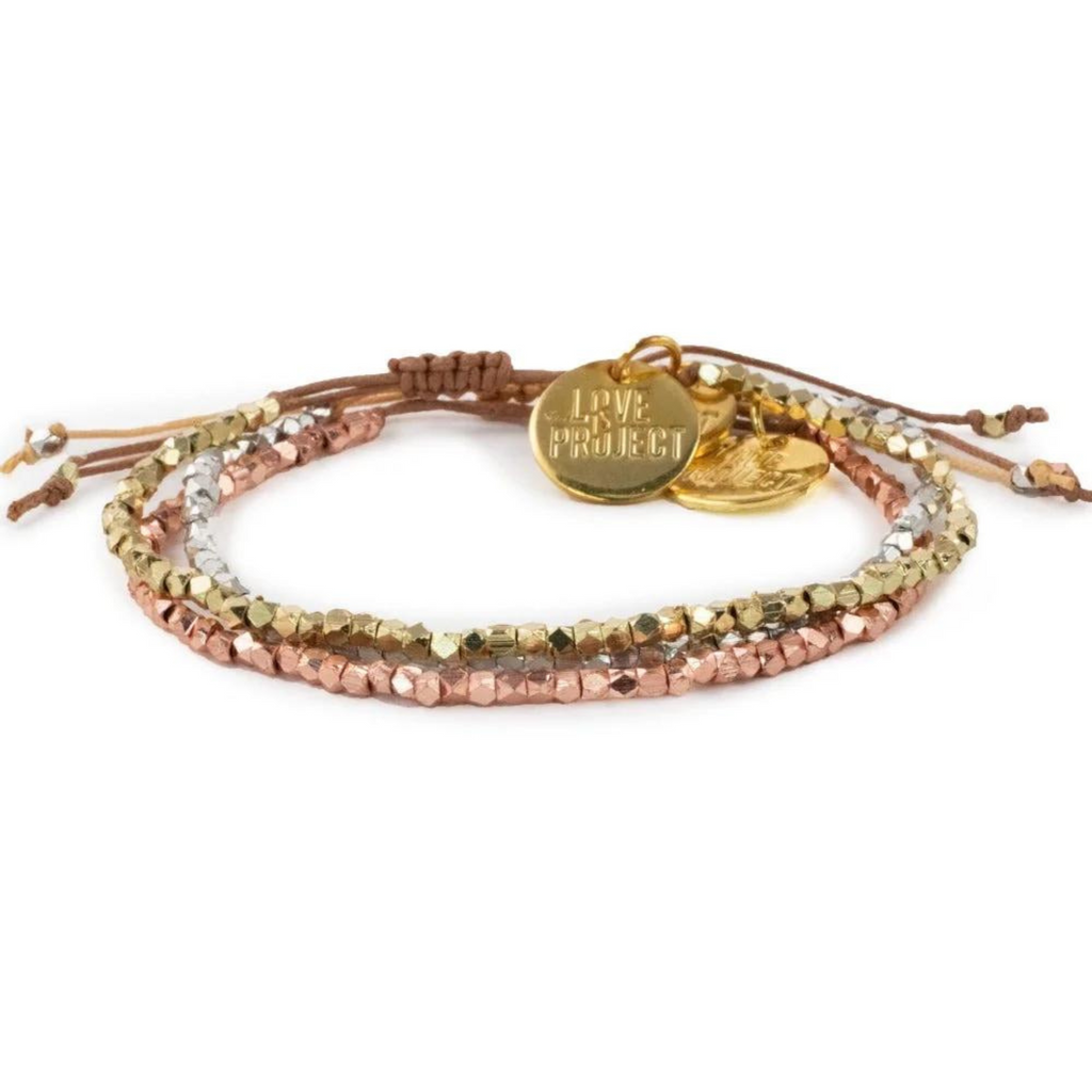 Product photography of a multi-strand bracelet featuring gold silver and rose gold faceted beads with a round "LOVE PROJECT" charm and a heart charm on an adjustable brown cord against a white background
