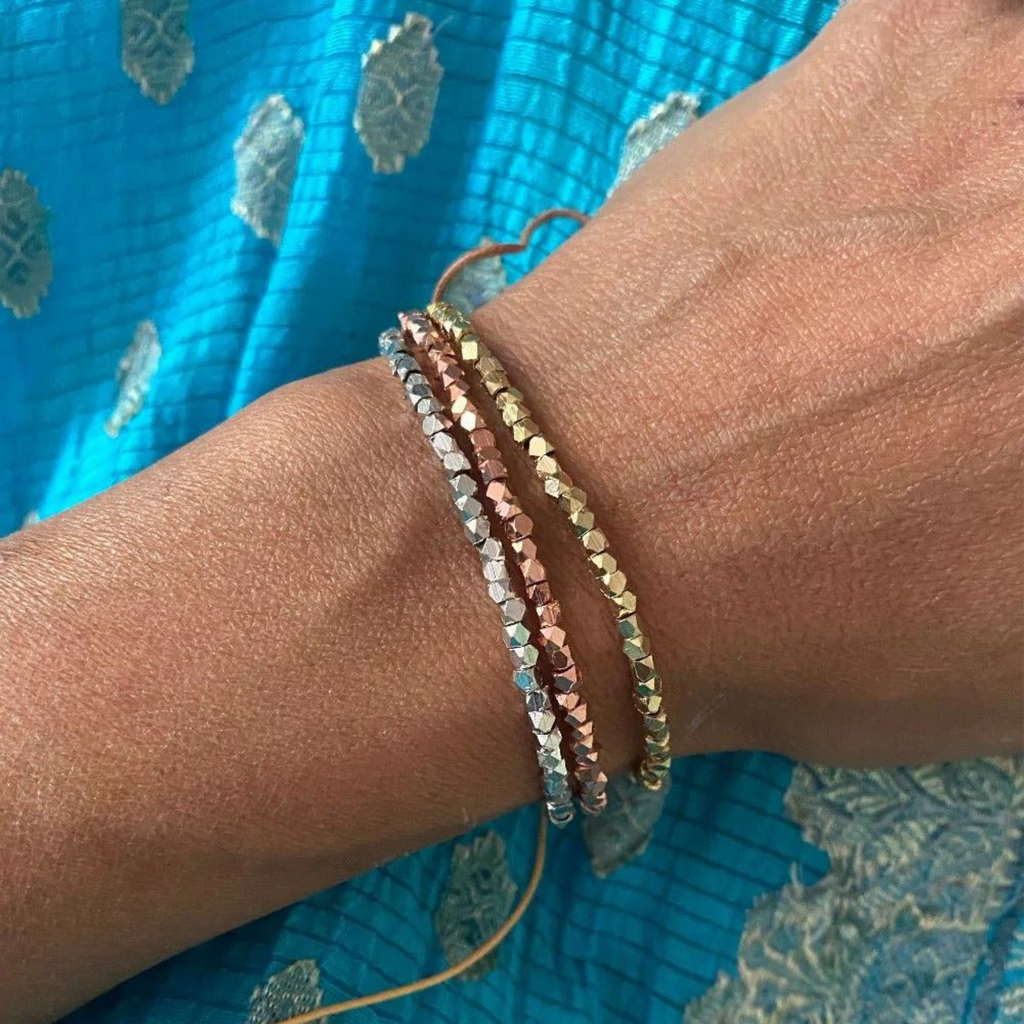 Close-up of a human wrist wearing three faceted hexagonal beaded bracelets in silver, rose gold, and gold, set against a bright blue patterned textile background with a thin brown string