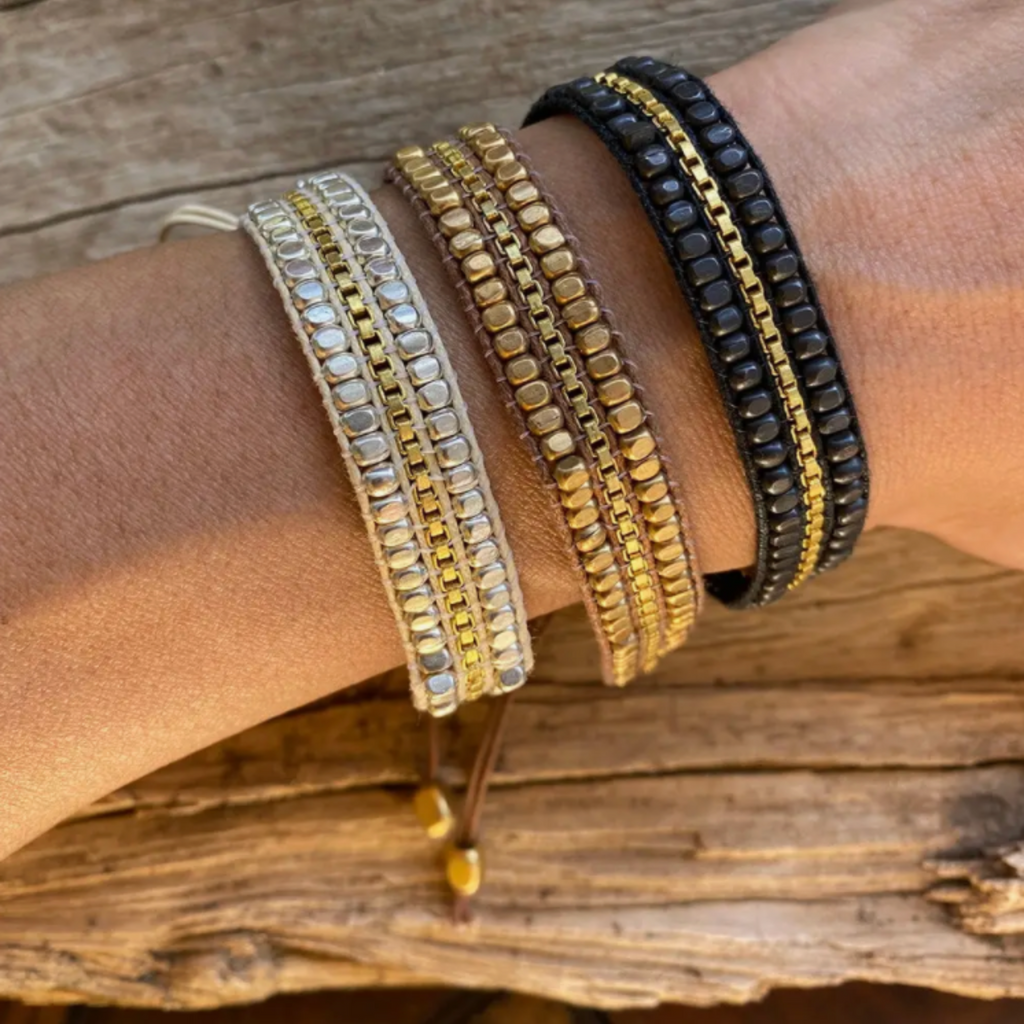Three beaded wrap bracelets are displayed on a person's wrist against a rustic wooden surface, featuring silver-toned, gold-toned, and black-beaded designs, each intricately interwoven with a golden chain