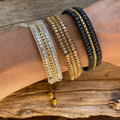 Three beaded wrap bracelets are displayed on a person's wrist against a rustic wooden surface, featuring silver-toned, gold-toned, and black-beaded designs, each intricately interwoven with a golden chain