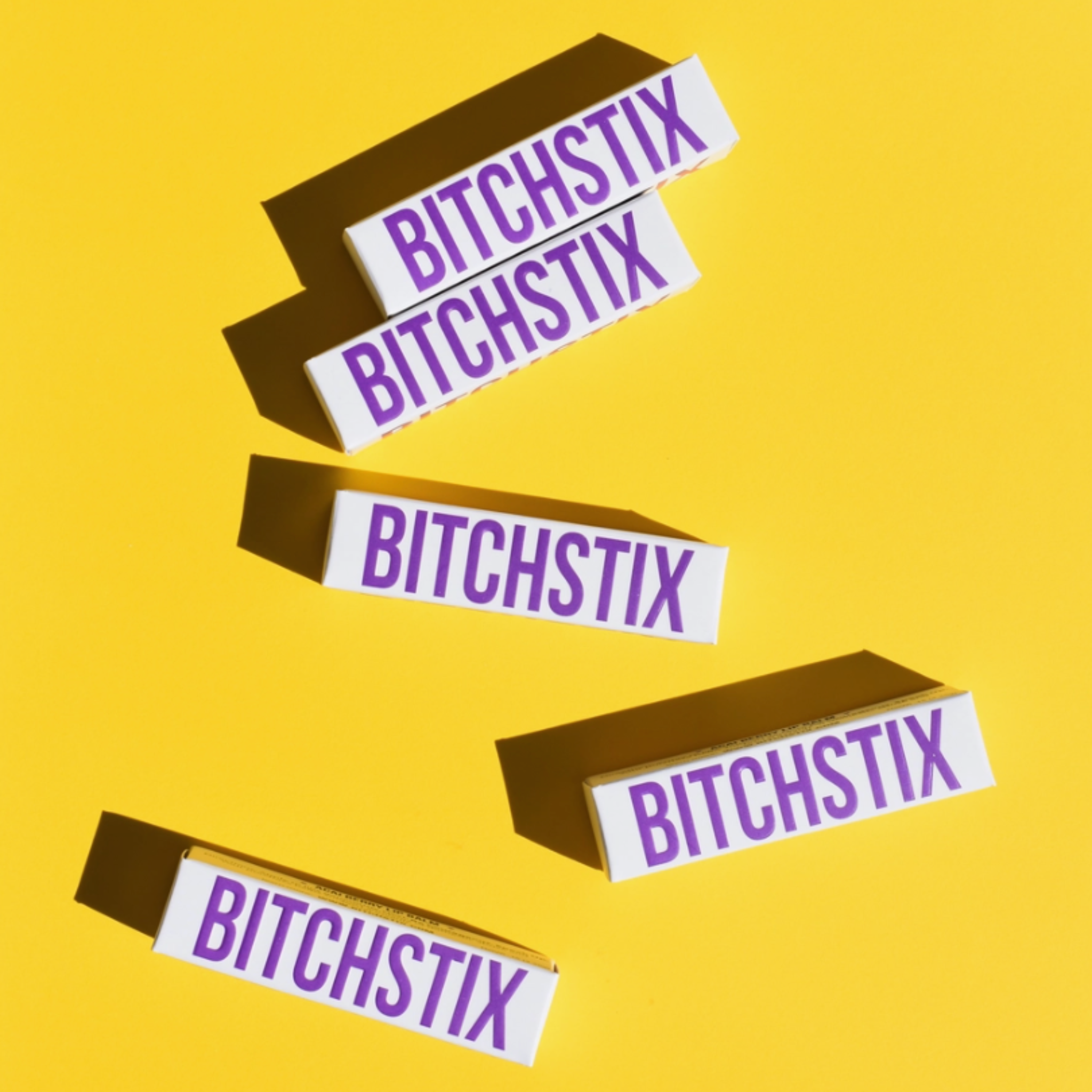 Product photography of five white BITCHSTIX lip balm boxes with bold purple lettering scattered dynamically on a vibrant yellow background, casting strong, sharp shadows